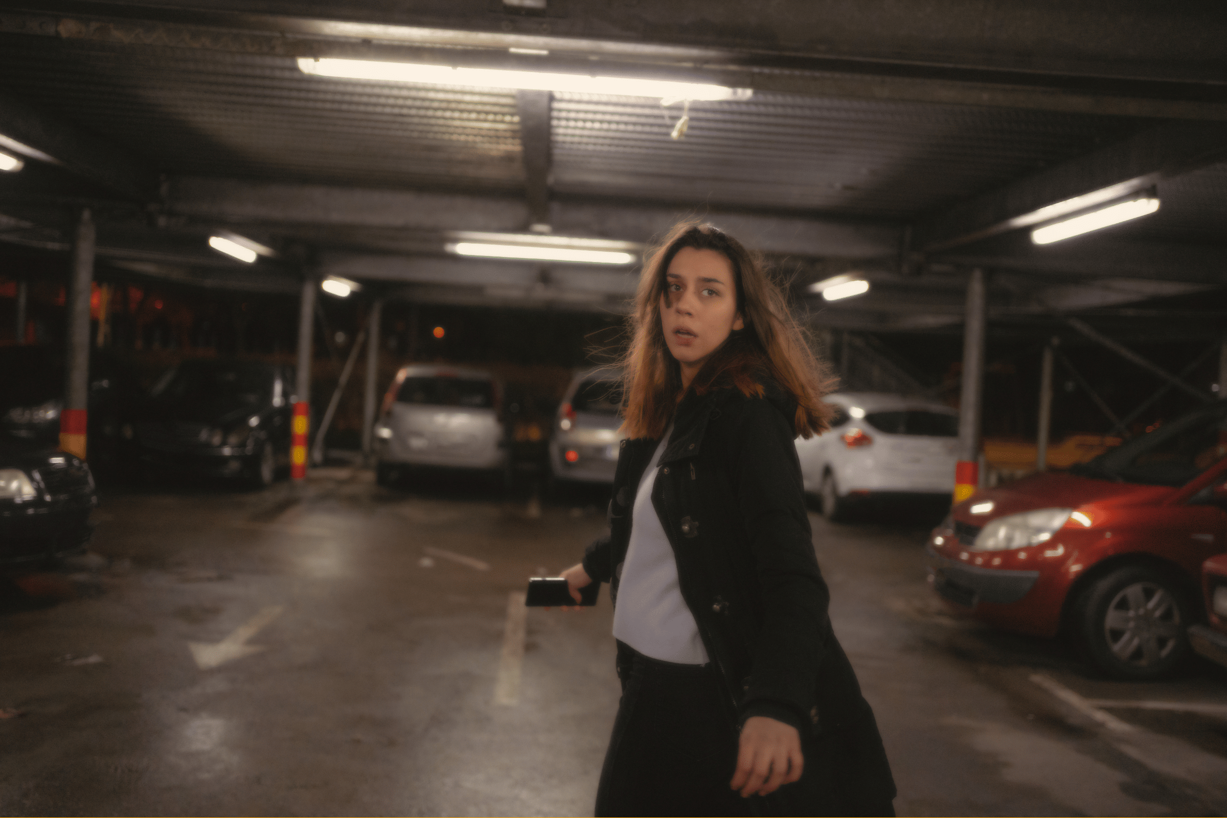 Woman in a dark jacket walking in a dim parking garage, looking back over her shoulder.
