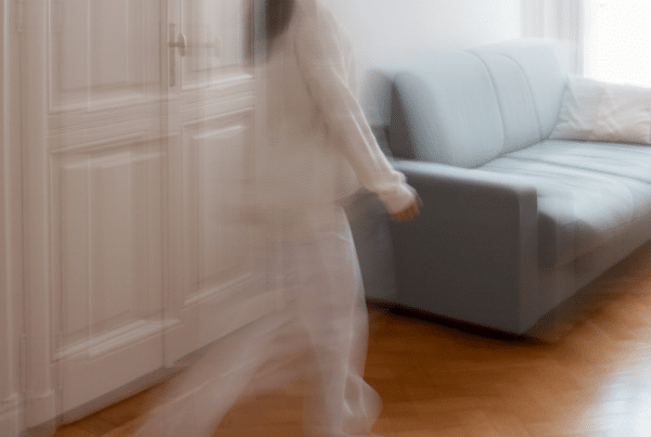 Motion-blurred person in a beige coat walking past a white paneled door toward a light-blue sofa in a bright living room area.