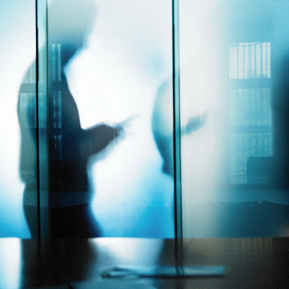Two blurred silhouettes behind frosted glass panels in a cool blue office setting.